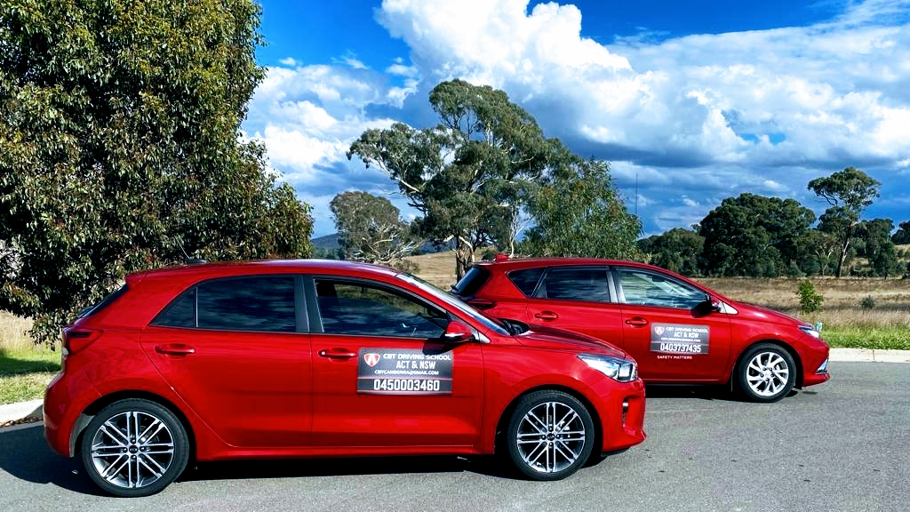 Two red driving school cars parked beside a tree on a country road.
